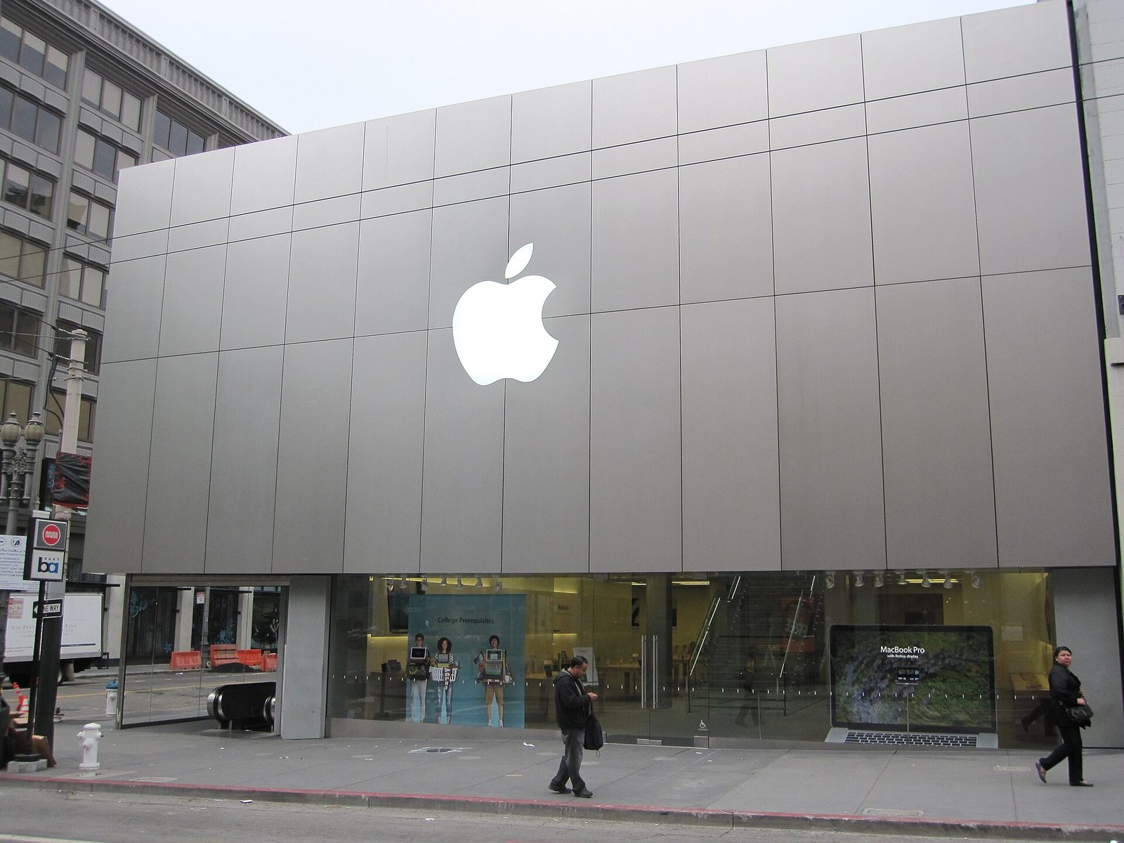 The exterior of an Apple Store in San Francisco: the company's white logo mounted on a grey panelled facade above ground-floor display windows, with pedestrians passing on the pavement