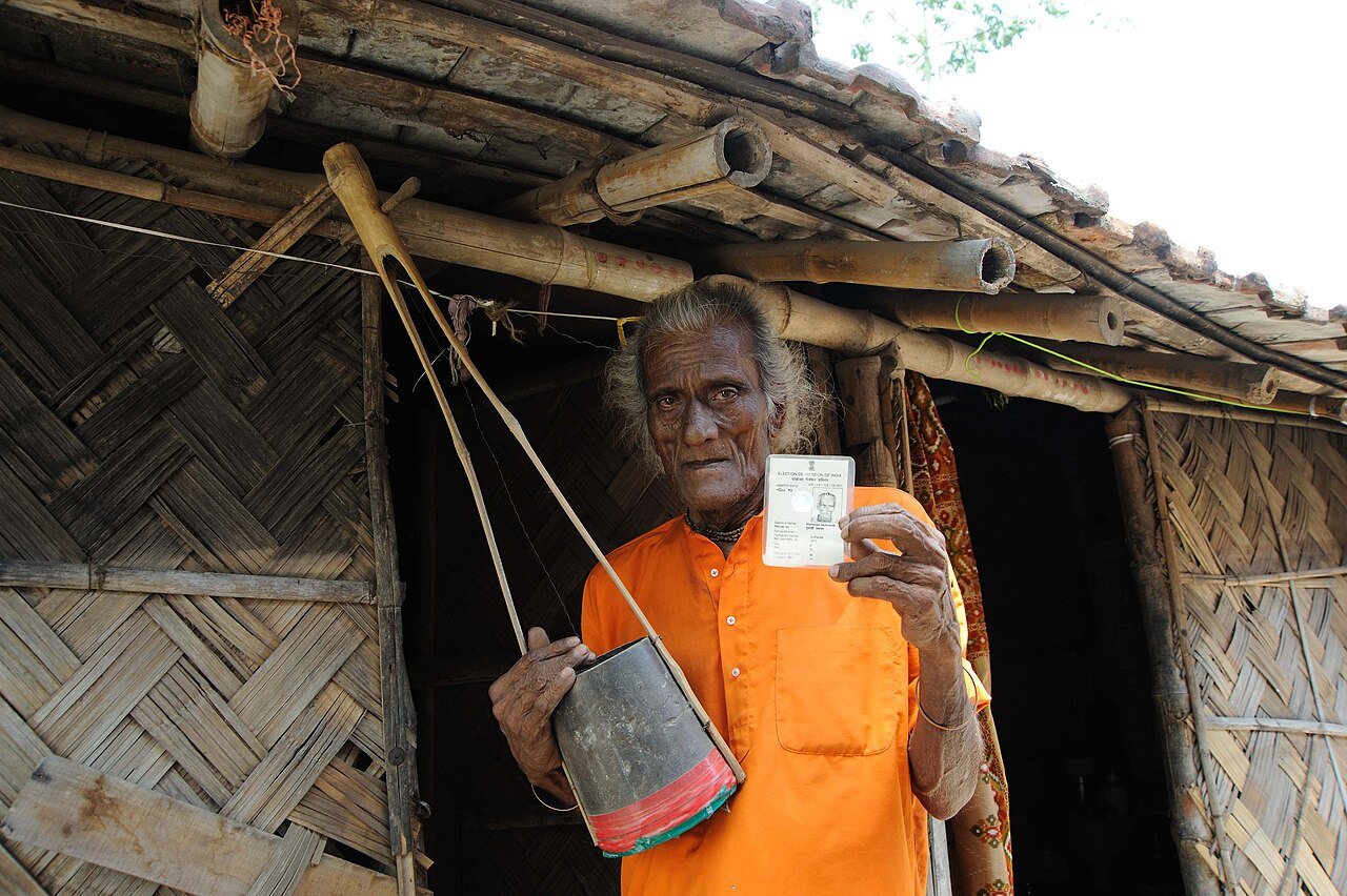 A voter holds up an Election Commission of India identity card outside a bamboo house